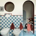 Modrobiely šachovnicový obklad, biela sanita, tehlovočervená linka a vysoký oblúkový priechod do vedľajšej miestnosti.