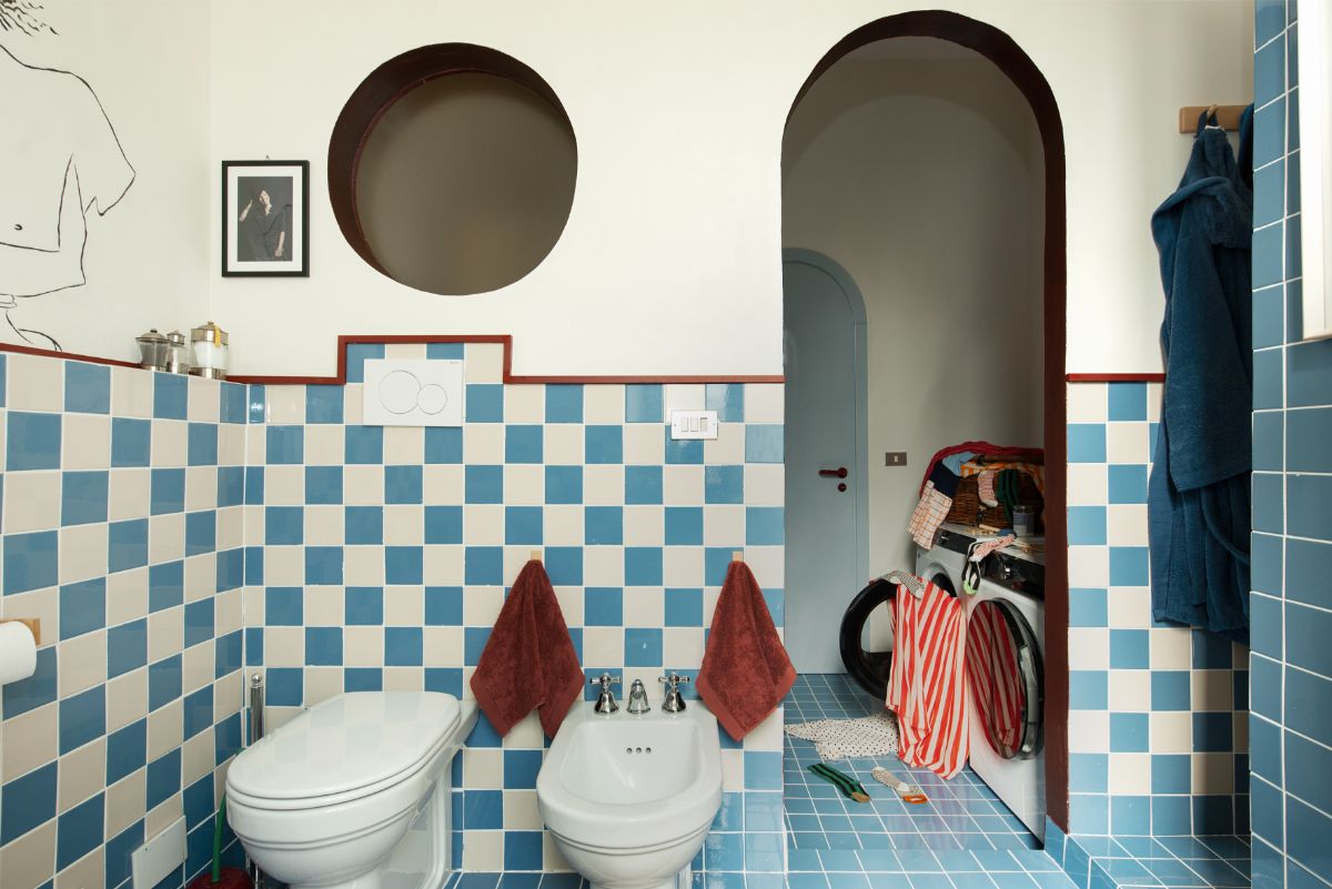 Modrobiely šachovnicový obklad, biela sanita, tehlovočervená linka a vysoký oblúkový priechod do vedľajšej miestnosti.