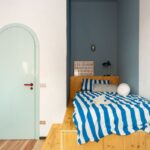 Modrý spací výklenok, vstavaná drevená posteľ so schodíkmi, modrobiely pruhovaný paplón a svetlomodré klenbové dvere.