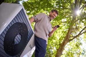 Muž stojí pod stromom pri tepelnom čerpadle a díva sa do mobilu.