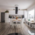 Moderná biela kuchyňa s čiernym digestorom, drevenou podlahou a dizajnovou lavicou pod oknom.