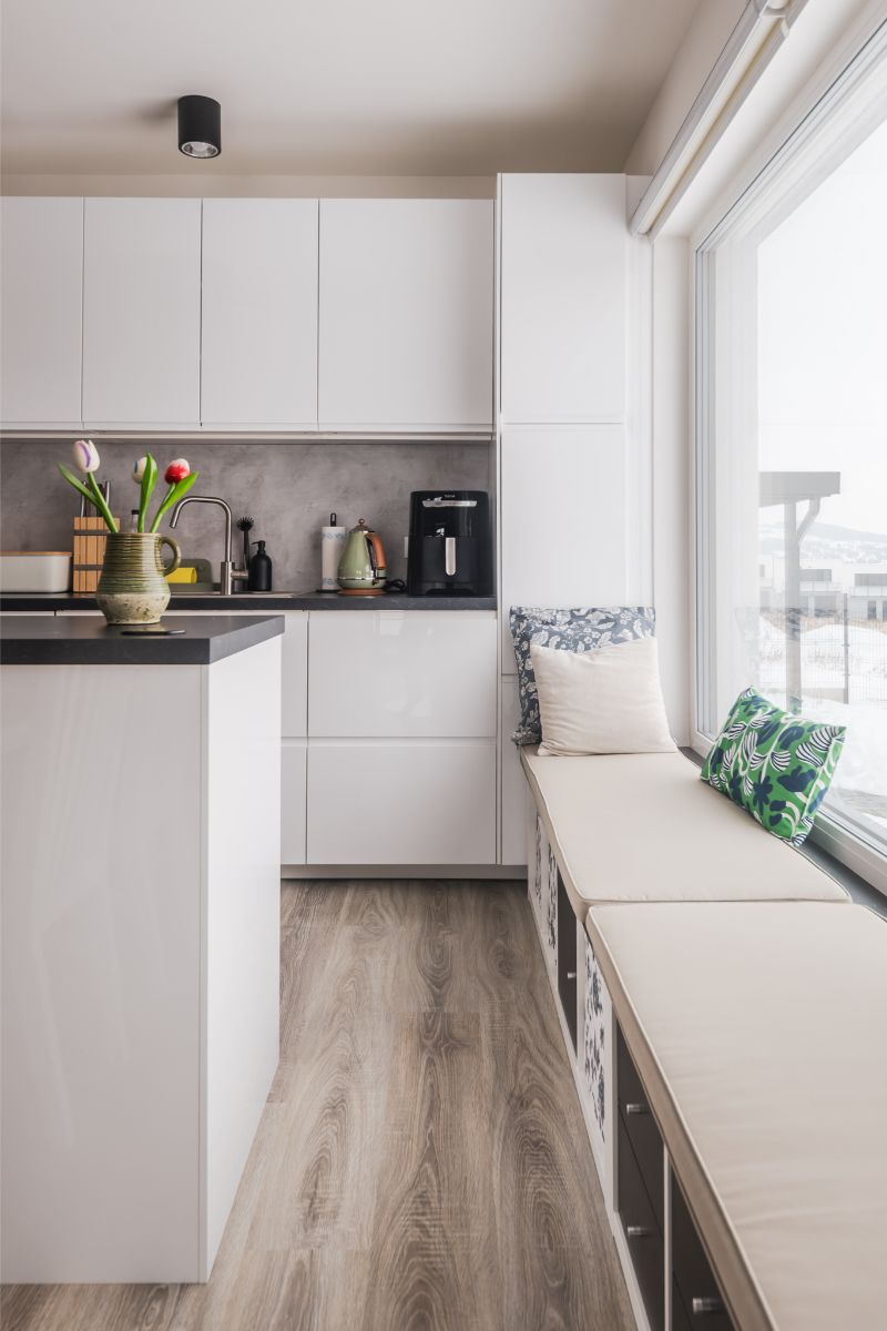 Minimalistická biela kuchynská linka a ostrovček s tmavou pracovnou doskou a moderným kávovarom.