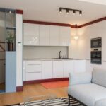 Kuchyňa s modernými bielymi skrinkami, vínovým soklom, sivou sedačkou a výrazným kobercom na podlahe.