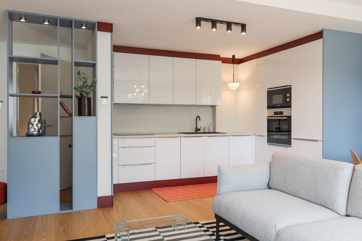 Kuchyňa s modernými bielymi skrinkami, vínovým soklom, sivou sedačkou a výrazným kobercom na podlahe.