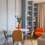 Jedálenský stôl s drevenými stoličkami, modrou knižnicou a výrazným oranžovým kreslom v rohu.