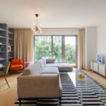 Obývacia izba so sivou rohovou sedačkou, pruhovaným kobercom, presklenými dverami na balkón a dizajnovým lustrom.