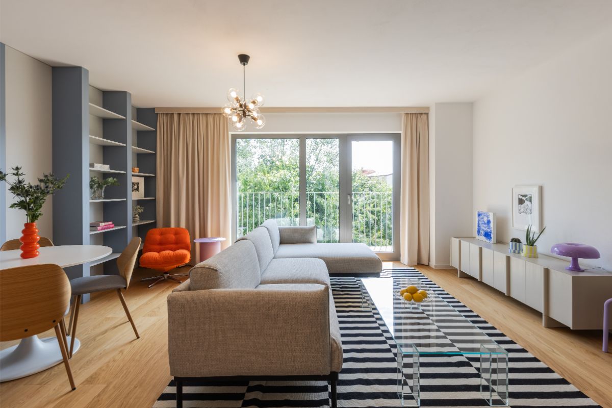 Obývacia izba so sivou rohovou sedačkou, pruhovaným kobercom, presklenými dverami na balkón a dizajnovým lustrom.