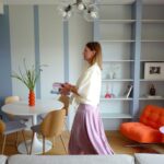 Obývacia izba s modrou pruhovanou stenou, bielou knižnicou, oranžovým kreslom a okrúhlym jedálenským stolom.