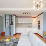 Kuchyňa spojená s obývačkou, bielou linkou, sivou sedačkou, čierno-bielym kobercom a modrými nábytkovými prvkami.