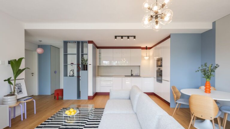 Kuchyňa spojená s obývačkou, bielou linkou, sivou sedačkou, čierno-bielym kobercom a modrými nábytkovými prvkami.