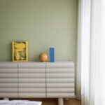 Spálňa so zelenou stenou, krémovou vlnitou komodou, modrou vázou a posteľou s fialovými pruhovanými obliečkami.