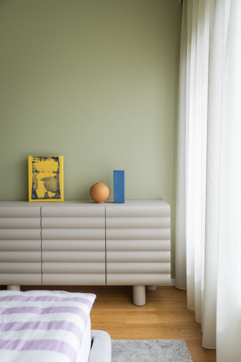 Spálňa so zelenou stenou, krémovou vlnitou komodou, modrou vázou a posteľou s fialovými pruhovanými obliečkami.