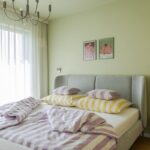 Spálňa so svetlozelenou stenou, sivou čalúnenou posteľou, fialovými pruhovanými obliečkami a moderným kovovým lustrom.