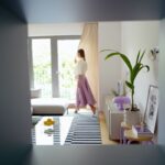 Obývacia izba s čiernobielym pruhovaným kobercom, bielou komodou, fialovou lampou a zelenou rastlinou v hlinenom kvetináči.