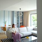 Obývacia izba so sivou rohovou sedačkou, skleneným konferenčným stolíkom, modrou knižnicou a bielym jedálenským stolom.