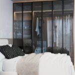 Manželská posteľ s vankúšmi, hnedou dekou a čiernymi stolíkmi pred veľkou sklenenou šatníkovou skriňou s kovovým rámom.