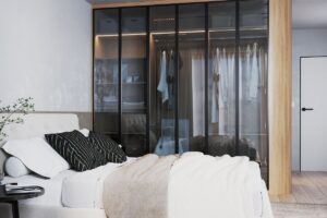 Manželská posteľ s vankúšmi, hnedou dekou a čiernymi stolíkmi pred veľkou sklenenou šatníkovou skriňou s kovovým rámom.