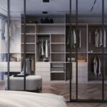 Moderná spálňa s bielou posteľou a šatníkovou skriňou s presklenými dverami a drevenými policami, v ktorej sú zavesené šaty a bundy.