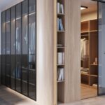 Rohová drevená šatníková skriňa so sklenenými dverami a osvetlenými policami, v ktorej sú uložené knihy, oblečenie a kabelka.