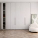 Veľká biela šatníková skriňa s otvorenými policami, v ktorej sú uložené oblečenie, topánky a doplnky, v spálni s bielymi stenami.