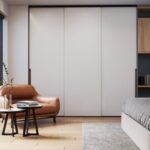 Moderná obývacia izba s béžovým kreslom, stolíkmi, rastlinami a bielou šatníkovou skriňou s posuvnými dverami a drevenými policami.