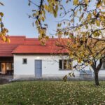 Pohľad zozadu na dedinský dom s červenou strechou, bielou a tehlovou fasádou, drevenou terasou a ovocným stromom.