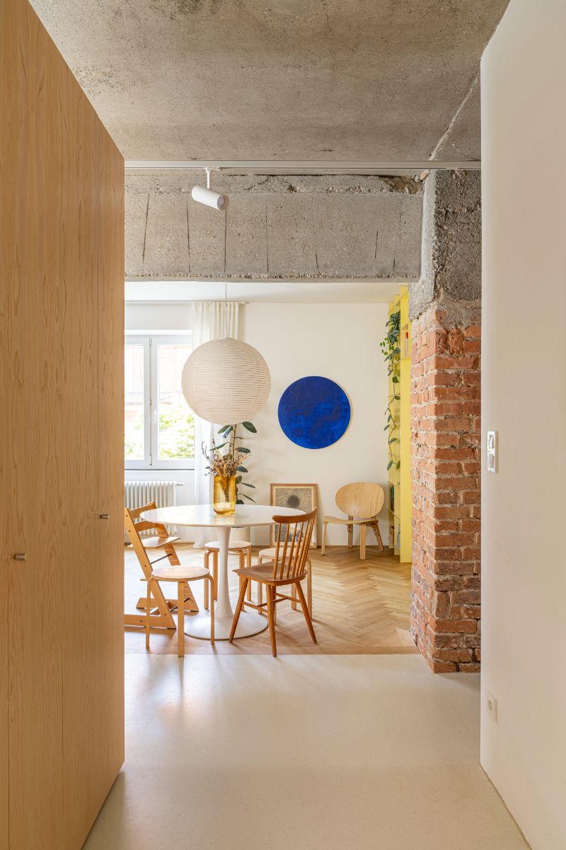 Jedáleň s bielym kruhovým stolom, drevenými stoličkami, priznaným betónovým stropom a červeným tehlovým stĺpom.