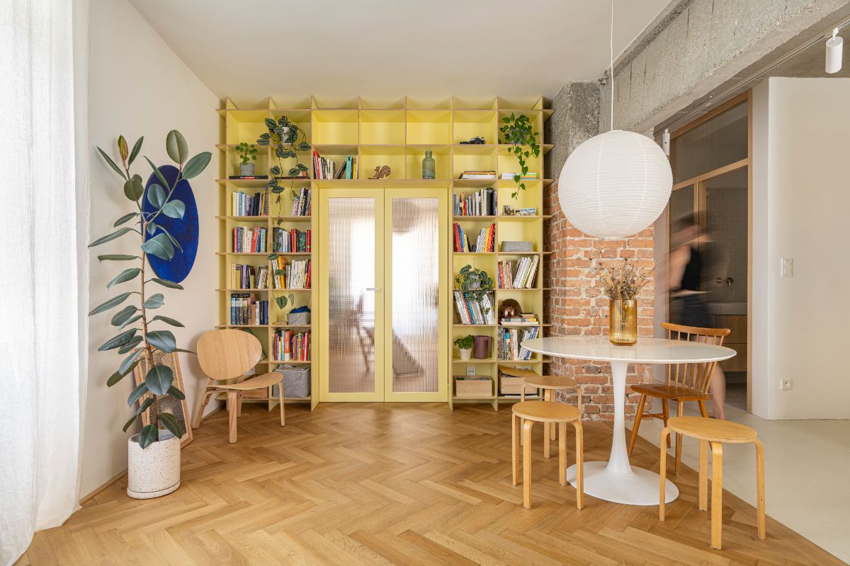 Jedáleň so žltou policovou stenou, bielym jedálenským stolom, drevenými taburetkami a parketami so vzorom stromčeka.