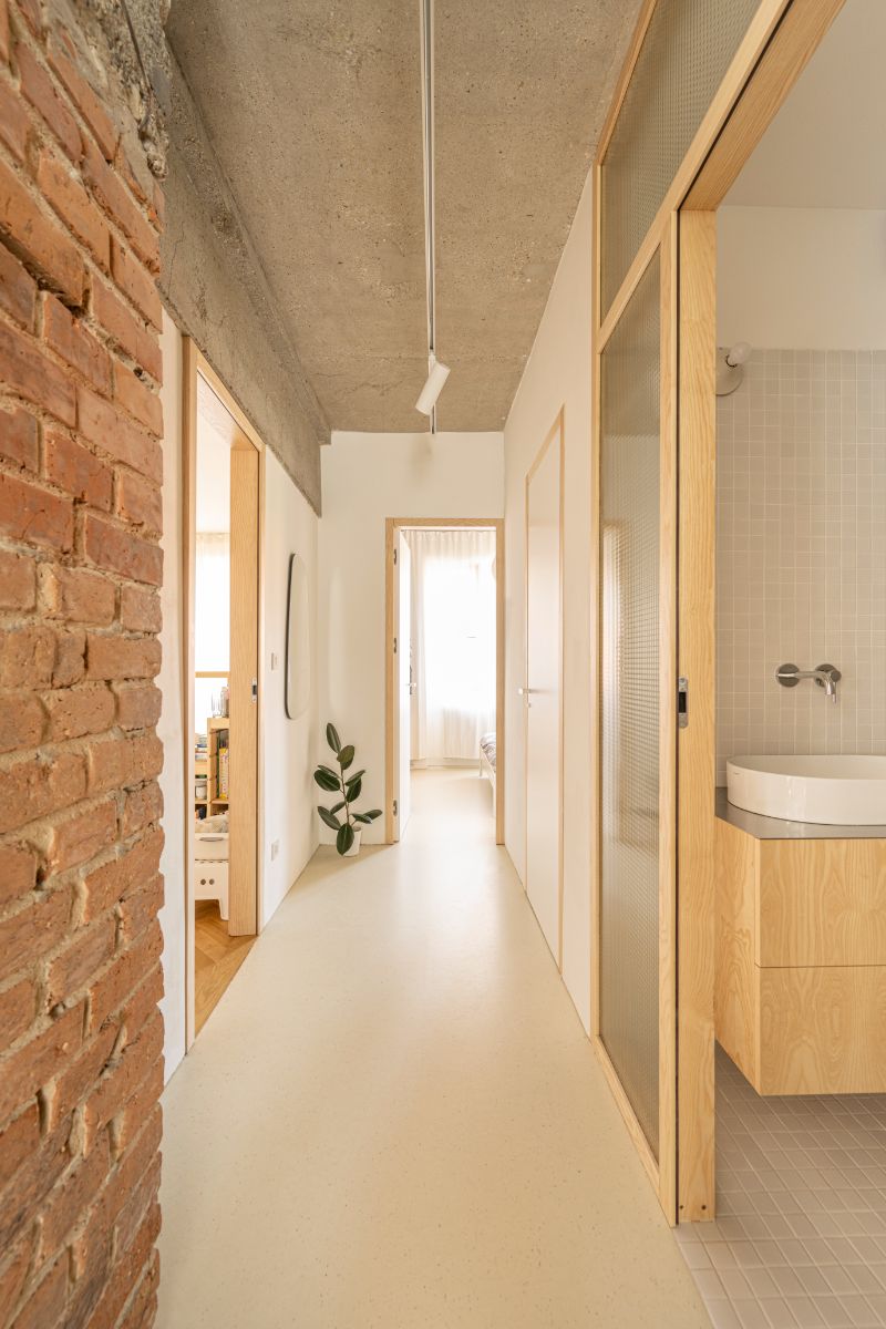 Chodba so svetlou podlahou, drevenými zárubňami, presklenými dverami do kúpeľne a surovým betónovým stropom.