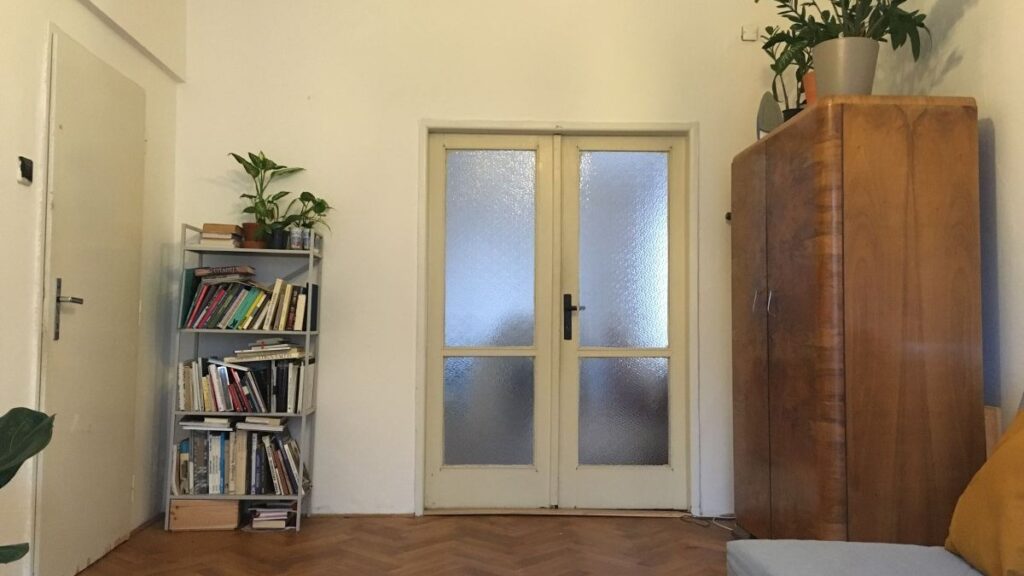 Pôvodný stav obývacej izby s bielymi stenami, retro skriňou, kovovým regálom a klasickými parketami.