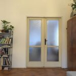 Pôvodný stav obývacej izby s bielymi stenami, retro skriňou, kovovým regálom a klasickými parketami.