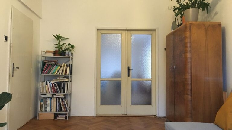 Pôvodný stav obývacej izby s bielymi stenami, retro skriňou, kovovým regálom a klasickými parketami.