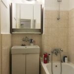 Kúpeľňa s béžovým keramickým obkladom, bielou skrinkou pod umývadlom, zrkadlovou skrinkou a malou vaňou.