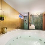 Kúpeľňa so zlatou lamelovou stenou, bielou vírivkou, zeleným vertikálnym obkladom a dizajnovým umývadlom na skrinke.