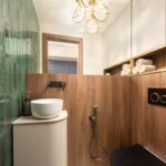 Kúpeľňa s dreveným stenovým panelom, zlatým WC, bielym oválnym umývadlom a visiacim guľovým svietidlom.