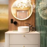 Kúpeľňa s bielou skrinkou, kruhovým podsvieteným zrkadlom, zeleným obkladom a čiernou vodovodnou batériou.