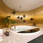 Kúpeľňa s kruhovou vírivkou, zlatou vlnitou stenou, visiacimi čiernymi kvetináčmi a terazzovou podlahou.