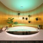 Kúpeľňa s bielou vírivkou, zlatou kovovou stenou, zeleným stropným osvetlením a sivou kamennou dlažbou.