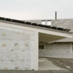 Prístavba so šikmou strechou, fotovoltickými panelmi, svetlým betónovým stĺpom a kamenným obkladom steny.