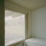 Spálňa s veľkým dreveným oknom, svetlou preglejkovou stenou a bielou posteľnou bielizňou.