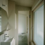 Chodba so svetlým dreveným obkladom, brúsenou betónovou podlahou a bielym keramickým umývadlom.