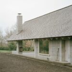 Exteriér domu so sivou omietkou, vysokým betónovým komínom a hnedou šindľovou strechou.