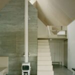 Obývacia izba so svetlým schodiskom, bielou pieckou a vysokou betónovou stenou.