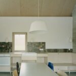 Kuchyňa s minimalistickým preglejkovým nábytkom, tmavým kamenným obkladom a visiacim bielym svietidlom.