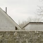 Kamenný plot pred sivou fasádou domu s hnedou strechou a kovovým komínom.