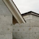 Fasáda domu zo sivého striekaného betónu s dreveným podhľadom a tmavou strešnou krytinou.