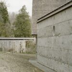 Vonkajšia stena budovy s drsnou betónovou omietkou a okrúhlym oknom vedľa dreveného plotu a lesa.