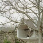 Biela budova s drevenou sedlovou strechou a stromom s holými vetvami.