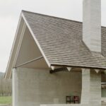 Detailnejší pohľad na rohovú časť domu s drsnou betónovou omietkou a sedlovou strechou s drevenými trámami.