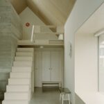 Vstupná chodba s preglejkovým schodiskom, vstavanou bielou skriňou a pohľadovým betónovým múrom.